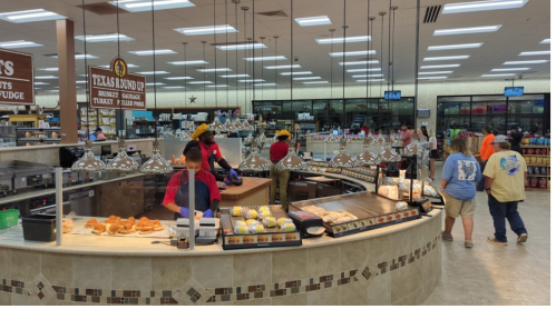 A busy food counter with staff serving various dishes in a well-lit store, featuring a variety of food options.