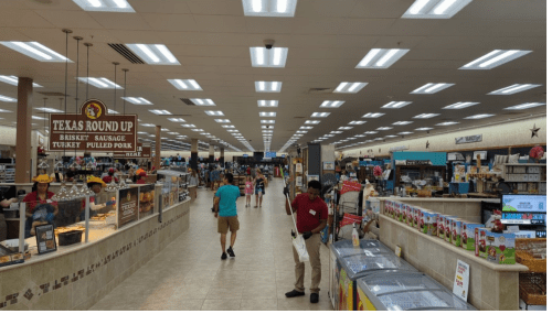 A spacious grocery store aisle with shoppers, displays of food, and bright overhead lights.