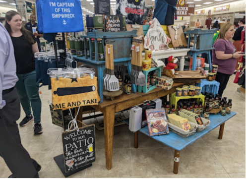 A display of kitchen items and food products in a store, with shoppers browsing nearby. Signs promote a patio theme.