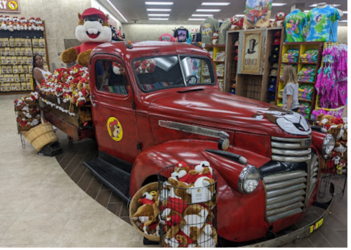 A vintage red truck filled with stuffed animals, set in a colorful store with various merchandise and shoppers.