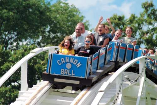A group of excited riders on the "Cannon Ball" roller coaster, enjoying a thrilling drop on a sunny day.