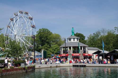 A bustling amusement park scene with a Ferris wheel, a pavilion, and crowds by a pond on a sunny day.