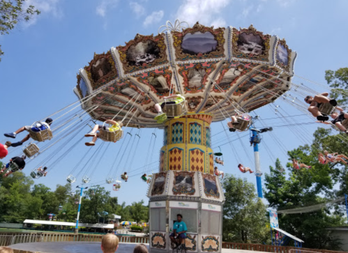 A colorful swing ride spins above a park, with riders in baskets enjoying the view on a sunny day.