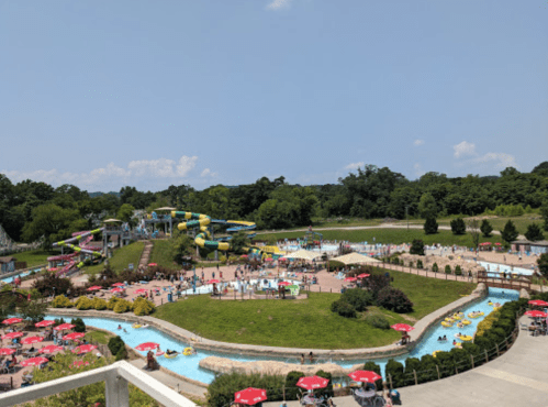 Aerial view of a water park with slides, lazy river, and sunbathers under red umbrellas on a sunny day.