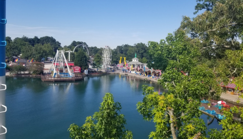 A scenic view of a amusement park by a lake, featuring rides, colorful tents, and lush greenery.