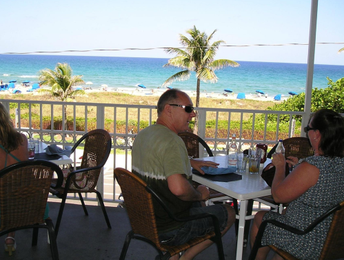 A sunny beach view from a restaurant patio with diners enjoying drinks and the ocean in the background.