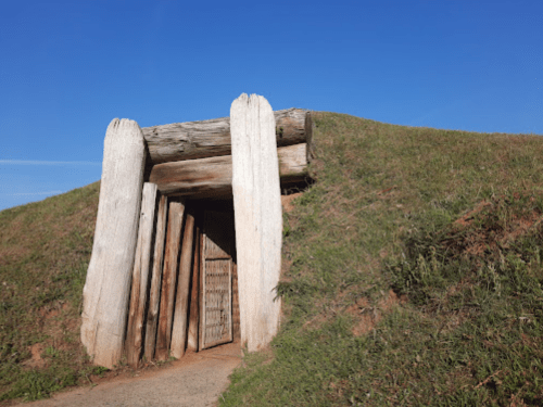 Ancient burial mound entrance with wooden beams, surrounded by green grass under a clear blue sky.
