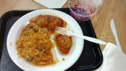 A plate with three meatballs in sauce, sauerkraut, and a cup of purple sauce on a tray. A plastic fork is on the plate.