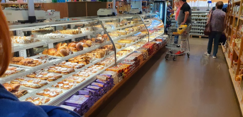 A bakery display filled with various pastries and baked goods, with shoppers browsing in a grocery store aisle.