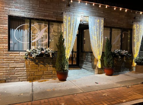 A warmly lit restaurant exterior with decorative curtains and potted plants, creating an inviting atmosphere at night.