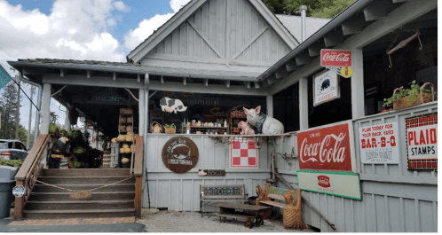 A rustic storefront with vintage signs, a pig statue, and outdoor seating under a blue sky with clouds.