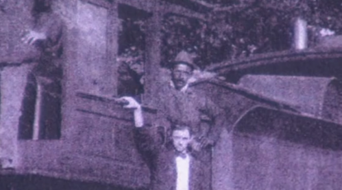 Two men pose on a vintage train, one standing with an arm raised, the other seated behind him.