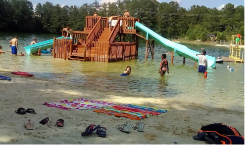 A sandy beach with people swimming and playing on a wooden structure with slides in the water. Colorful towels are spread out.
