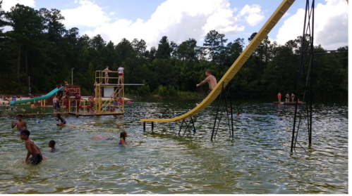 A sunny day at a lake with people swimming, a water slide, and a diving platform. Kids enjoy the water and slides.