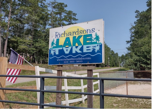 Sign for Richardson's Lake with an American flag nearby, surrounded by trees and a gravel road.