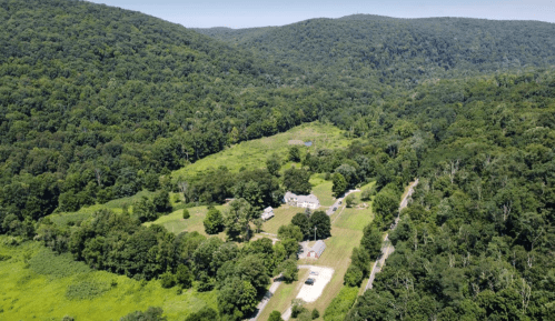 Aerial view of a lush green valley with a small farmhouse and surrounding trees, mountains in the background.
