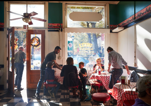A cozy diner scene with patrons at checkered tables, a server attending to a family, and sunlight streaming through the door.