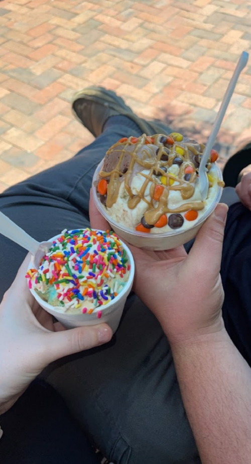 Two hands holding ice cream cups: one topped with colorful sprinkles and the other with peanut butter and candy.