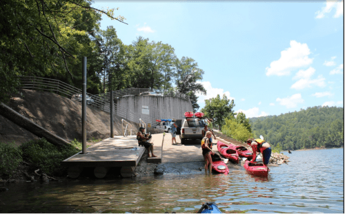 A group of people unload kayaks at a dock by a lake, with a dam and trees in the background under a clear blue sky.