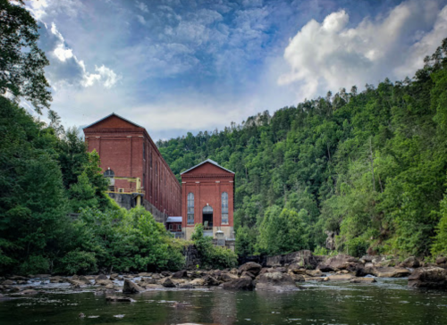 Historic red brick building nestled among lush green trees by a river, under a partly cloudy sky.