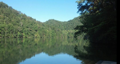 A serene lake surrounded by lush green mountains under a clear blue sky, reflecting the landscape on the water's surface.