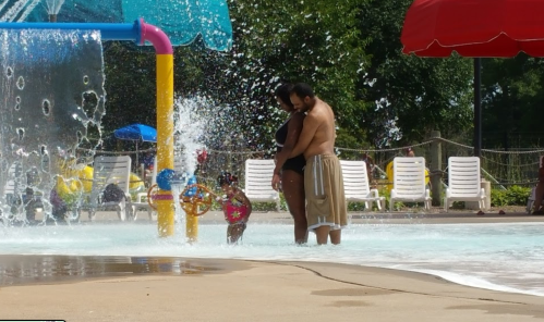 A couple embraces in a splash pad area while a child plays nearby, surrounded by water features and lounge chairs.