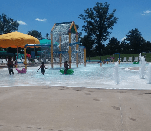 Children playing in a splash pad with water features, colorful slides, and sunny skies in the background.