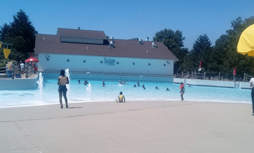 A sunny day at a pool with people swimming and playing, surrounded by trees and a building in the background.