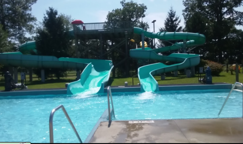 Two water slides beside a swimming pool, with a grassy area and trees in the background on a sunny day.