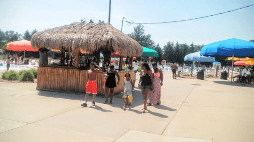 A family walks past a tiki bar at a water park, with colorful umbrellas and a pool in the background.