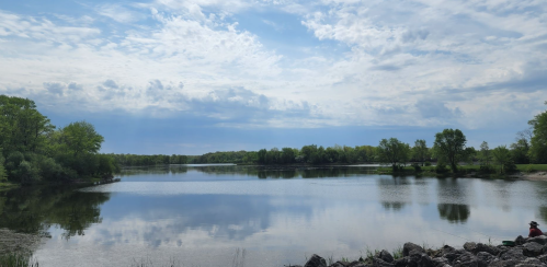 A serene lake surrounded by lush greenery under a partly cloudy sky, with calm water reflecting the landscape.