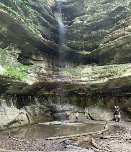 A group of people stands in a cave with a waterfall cascading down rocky walls, surrounded by lush greenery.