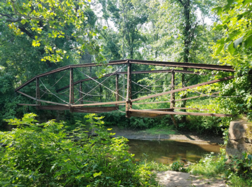 A rusted metal bridge spans a small creek, surrounded by lush green trees and foliage.