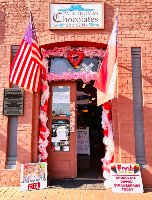 A charming chocolate shop entrance decorated with flags, hearts, and a sign for fresh chocolate-dipped strawberries.