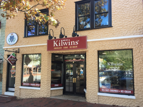 Exterior of a Kilwins store with a textured beige facade, large windows displaying chocolates and ice cream.
