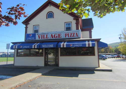 A two-story building with a sign reading "Village Pizza" and a blue awning, surrounded by trees and a parking lot.