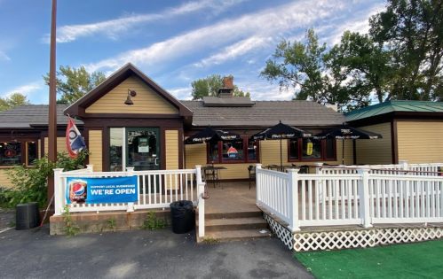 A cozy restaurant with outdoor seating, featuring umbrellas and a sign that says "We're Open." Trees in the background.