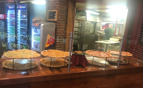 A display of various pizzas in a restaurant, with a kitchen visible in the background and drinks in vending machines.