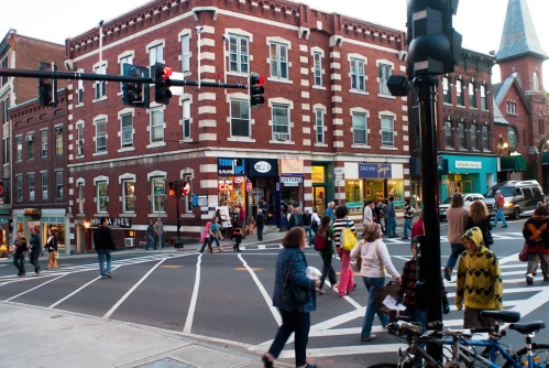 Busy street scene with pedestrians crossing, red traffic lights, and brick buildings in an urban setting.