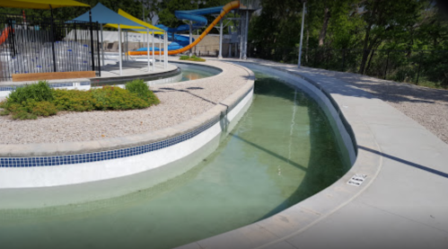 A winding lazy river pool with clear water, surrounded by landscaping and water slides in the background.