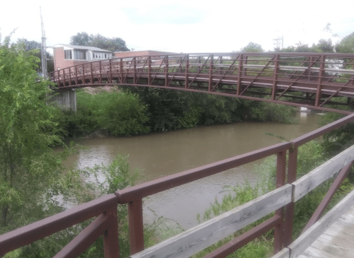 A metal footbridge arches over a muddy river, surrounded by greenery and a wooden walkway.