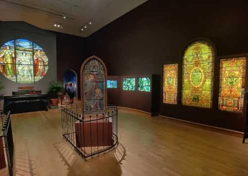 A gallery featuring stained glass windows, including a large circular piece and several colorful panels, with visitors in the background.