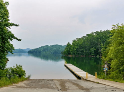 A calm lake with a wooden dock, surrounded by lush green trees and distant hills under a cloudy sky.