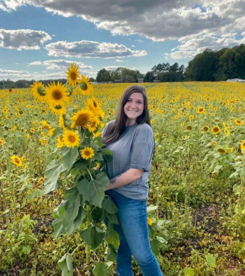 A smiling woman stands in a sunflower field, holding a large bouquet of sunflowers against a blue sky.