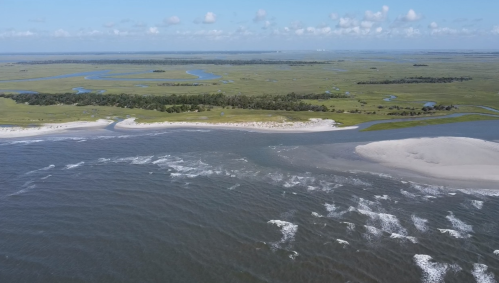 Aerial view of a coastal landscape with sandy shores, winding rivers, and lush green vegetation under a clear blue sky.