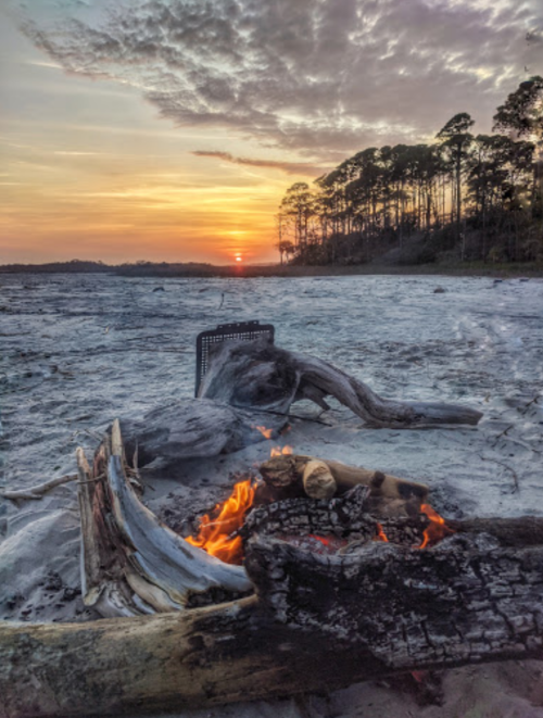 A serene sunset over a sandy beach with a small campfire and driftwood in the foreground.