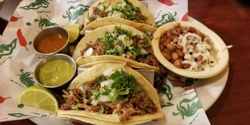 Three tacos filled with shredded meat, topped with cilantro, served with lime and two sauces, alongside a bowl of beans.