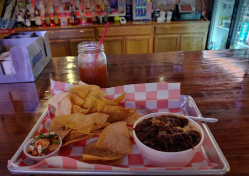 A tray with tortilla chips, salsa, and a bowl of chili on a wooden bar, with a drink in a jar in the background.
