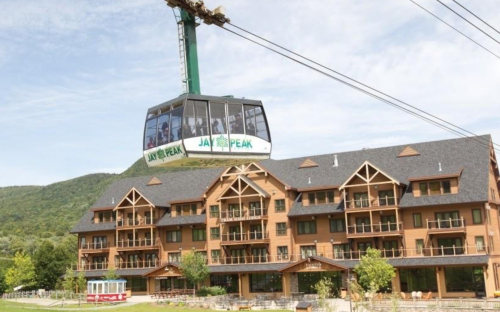 A gondola lifts above a large wooden lodge at Jay Peak, surrounded by green mountains and trees.