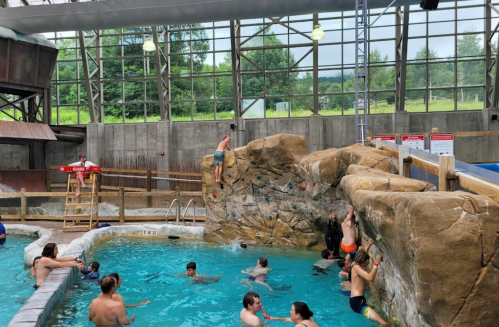 A lively indoor pool area with a rock climbing wall, kids swimming, and a lifeguard on duty.